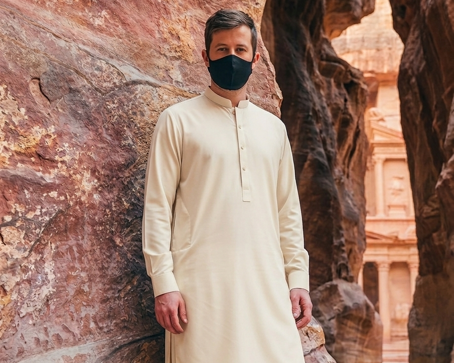 Man wearing a beige traditional outfit with a black mask standing against a rock wall.